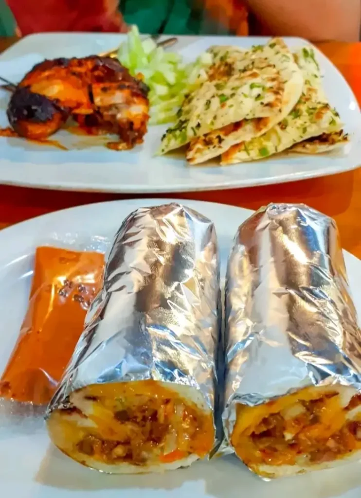 A close-up shot of Sadia's Kitchen's meal combo featuring a sliced chicken shawarma, a piece of chicken chap, and a folded butter naan on a white plate. The food is presented ready to eat.