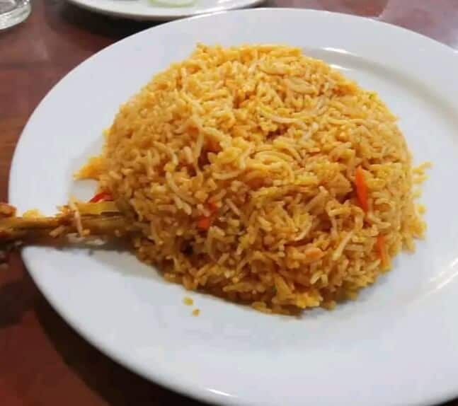 A close-up of a plate of Sadia's Kitchen's Chicken Biryani, showing a generous portion of aromatic rice and a large piece of chicken. The dish is garnished with fried onions and a side of salad.