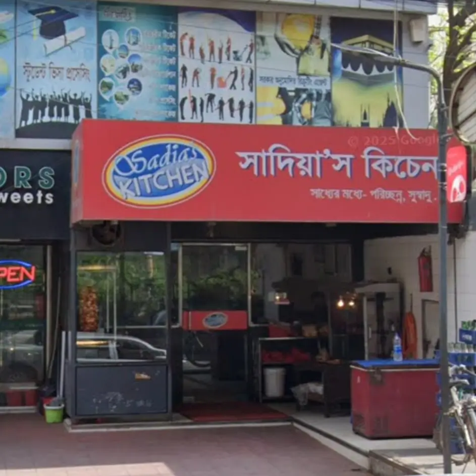 Exterior view of Sadia's Kitchen at Muradpur Circle, Chittagong, with the red and white sign clearly visible. The photo captures the busy street-level presence of the restaurant, popular for its shawarma and quick bites.